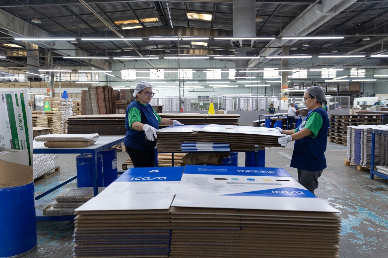 Operários separando folhas de papelão em um armazém com pilhas de caixas achatadas e equipamentos de transporte.