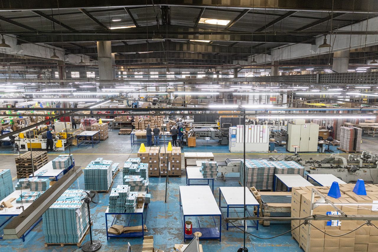 Interior de um grande armazém com pilhas de caixas, paletes e equipamentos industriais sob luzes fortes no teto.
