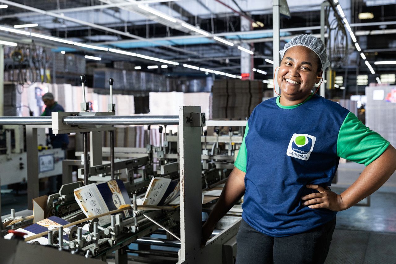 Operário sorridente de camisa azul em pé em uma fábrica movimentada ao lado de máquinas industriais.