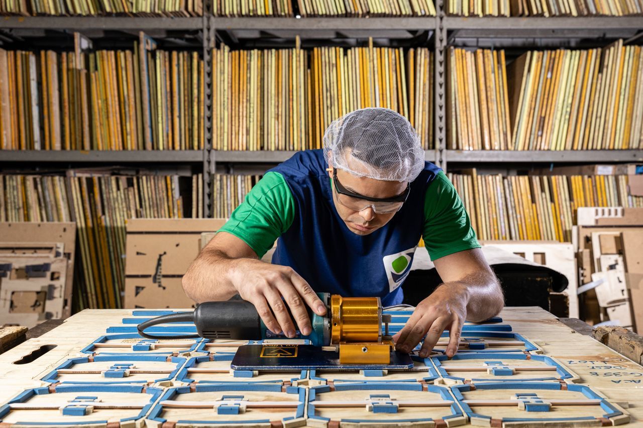 Pessoa montando uma tábua de madeira com grampos em uma oficina, com prateleiras de materiais ao fundo.