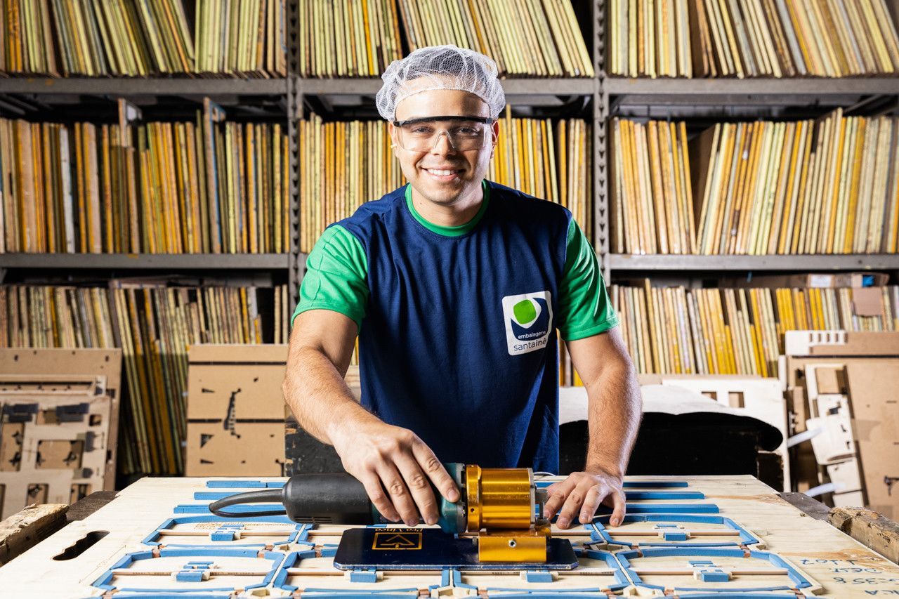 Homem sorrindo atrás de uma mesa com equipamentos eletrônicos, com prateleiras de discos ao fundo.