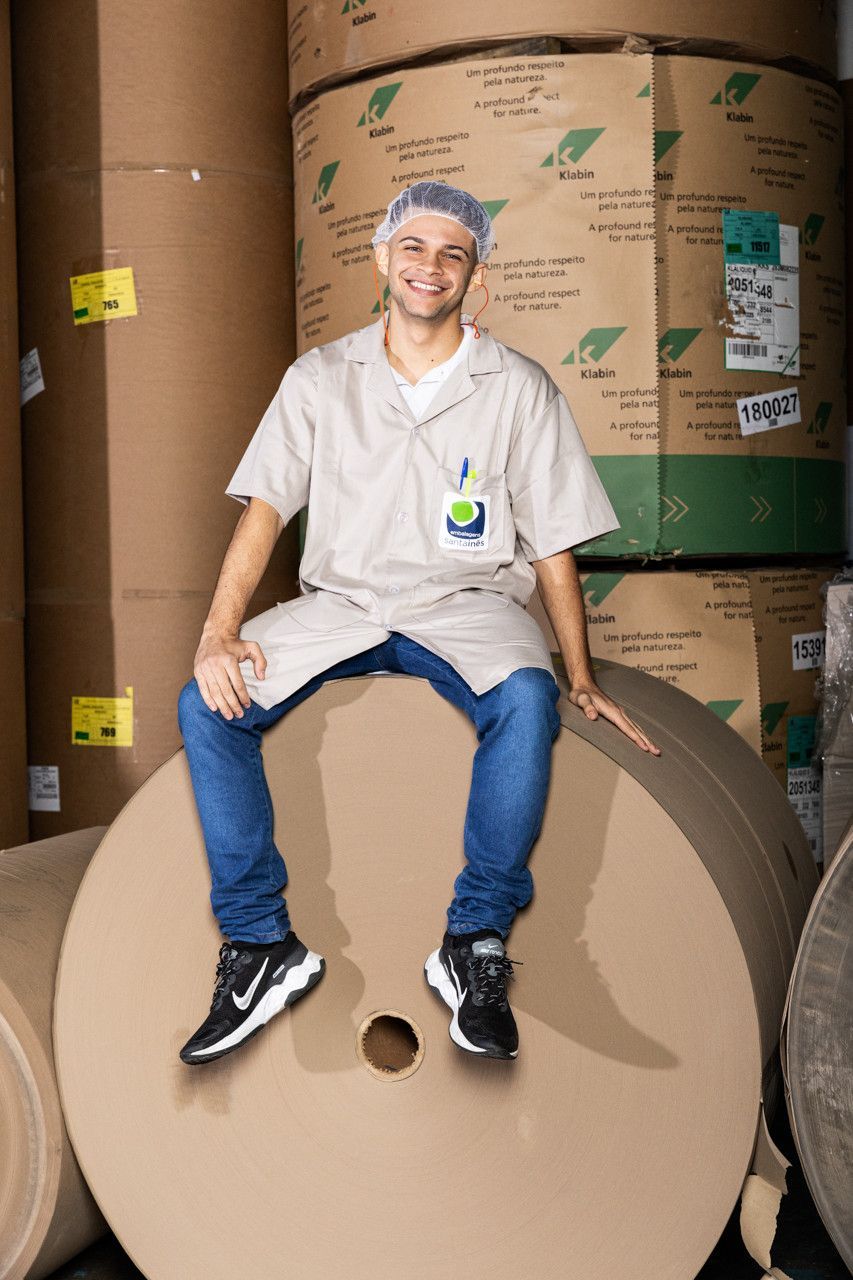 Pessoa vestindo camisa branca e calça jeans sentada em um grande rolo de papelão em um armazém.