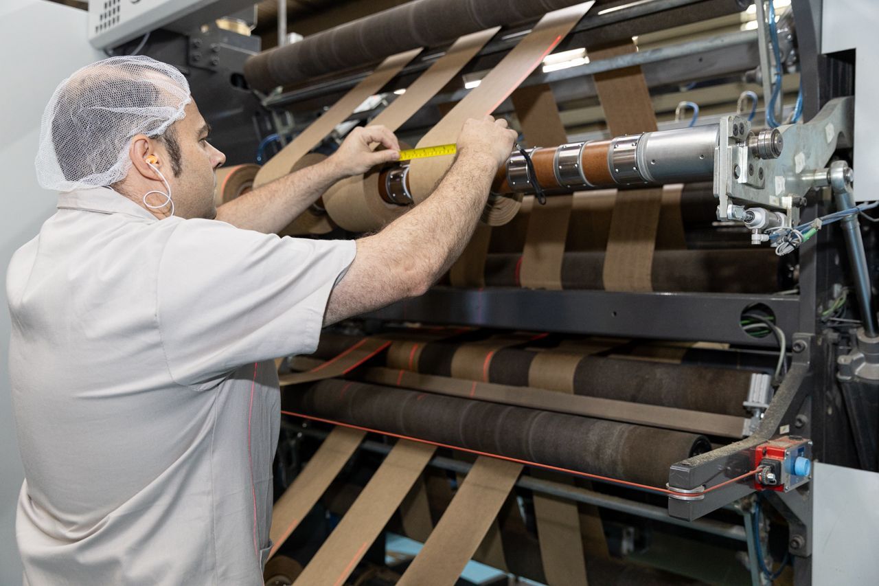 Operário trabalhando em uma máquina de impressão industrial em uma fábrica, ajustando os rolos e o material impresso.