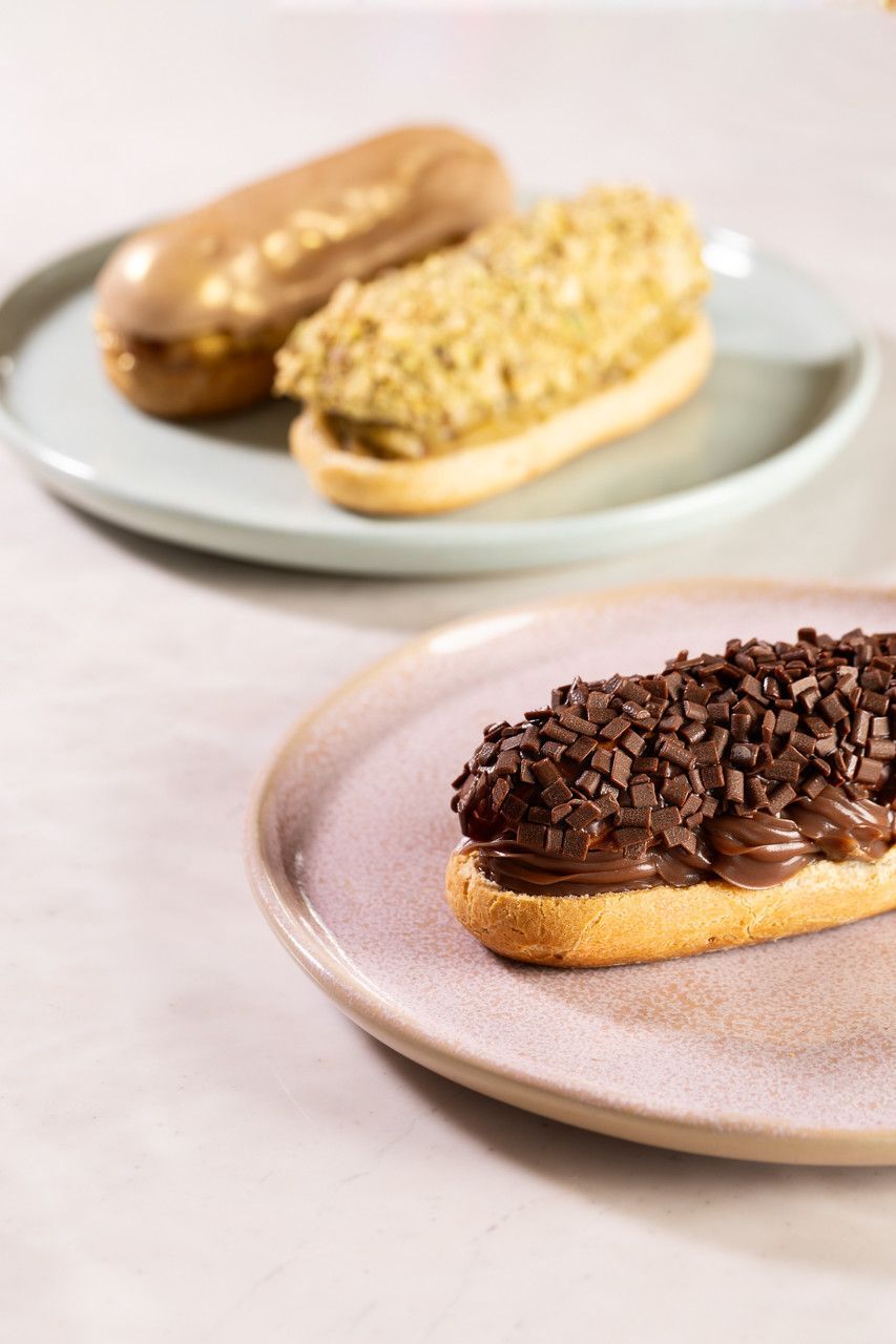 Três éclairs em pratos, cobertos com chocolate e nozes, sobre um fundo claro.