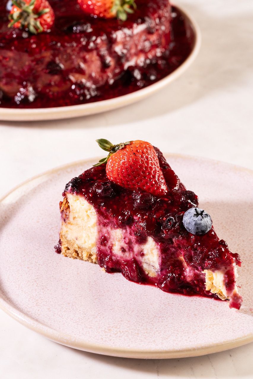 Fatia de cheesecake com cobertura de frutas vermelhas em um prato rosa claro, com um bolo inteiro desfocado ao fundo.