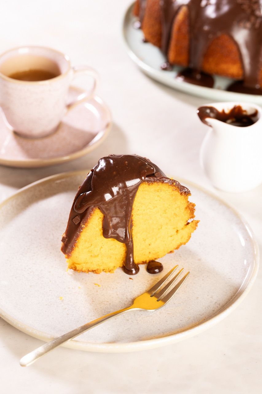 Fatia de bolo Bundt com cobertura de chocolate em um prato branco com um garfo dourado, ao lado de xícaras de café e um bolo inteiro.