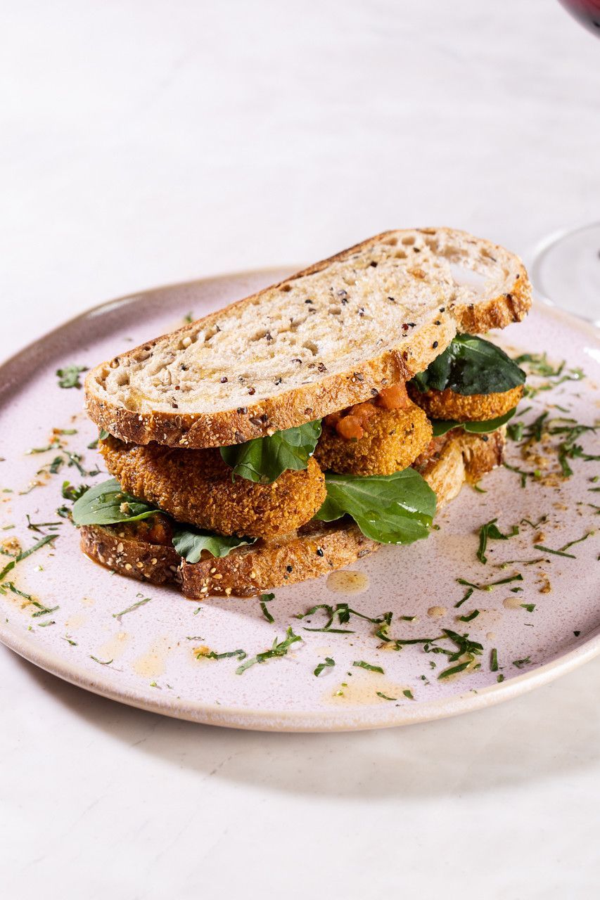Sanduíche aberto com recheio frito, verduras e ervas em um prato branco.