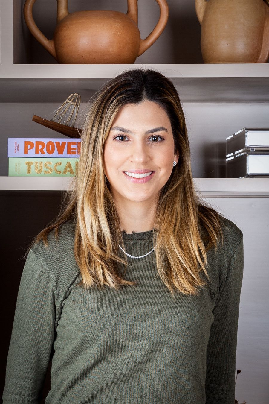 Mulher sorridente com um suéter verde-oliva em pé em frente a prateleiras com cerâmica e livros.