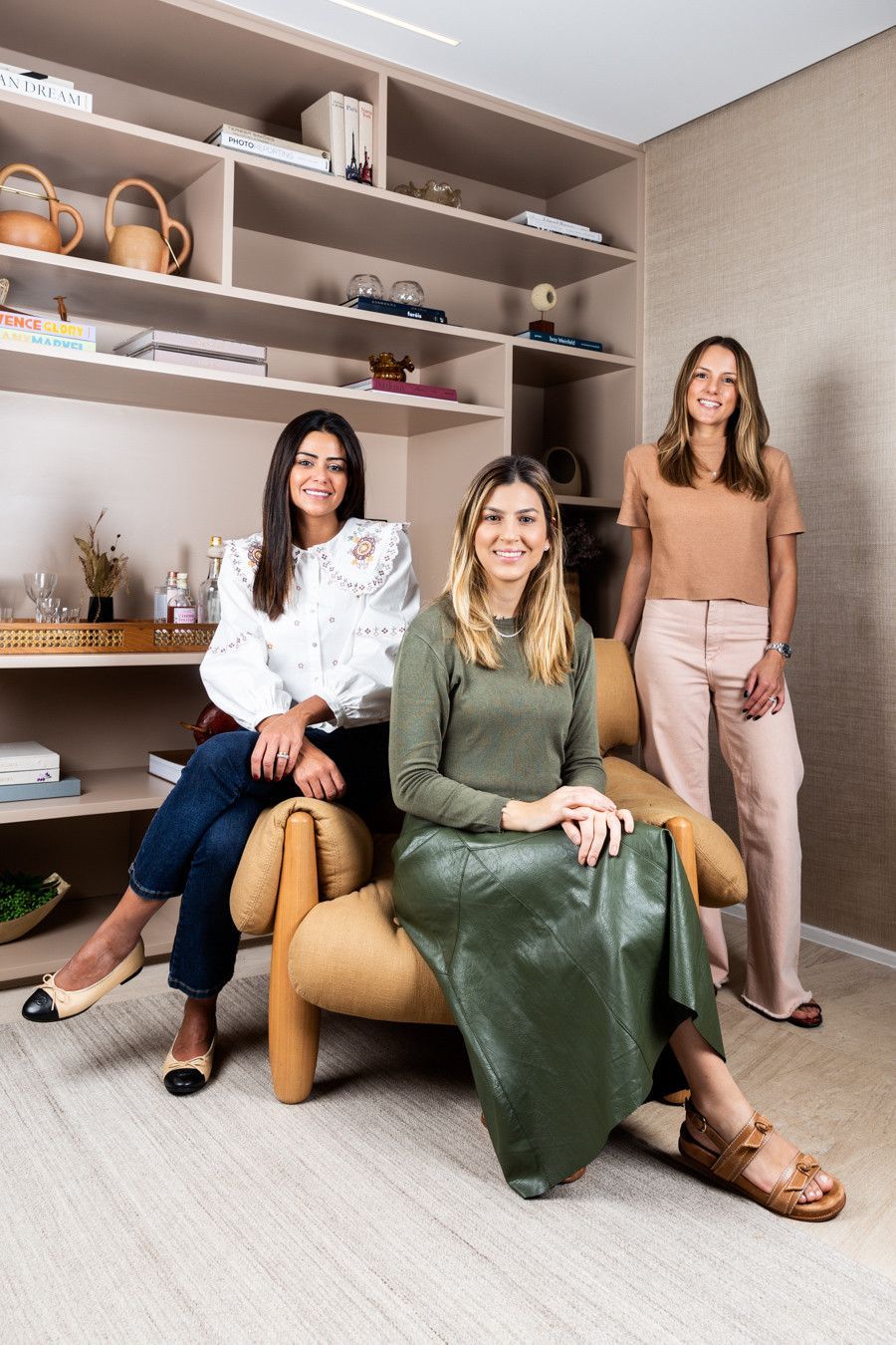 Três mulheres posando em uma sala de estar moderna com prateleiras e uma cadeira bege.