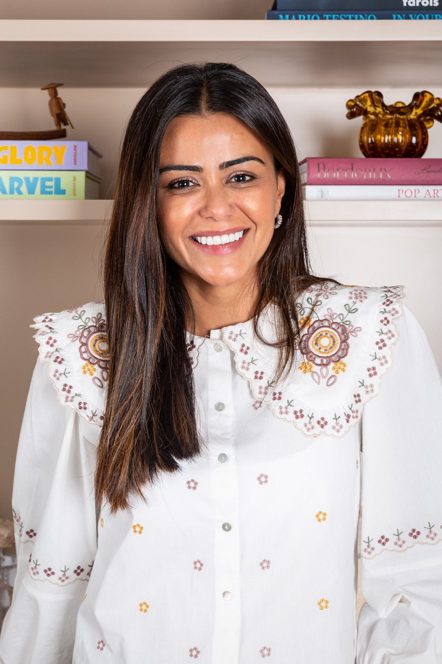 Pessoa sorridente vestindo uma blusa branca bordada em pé em frente a prateleiras com livros e objetos de decoração.