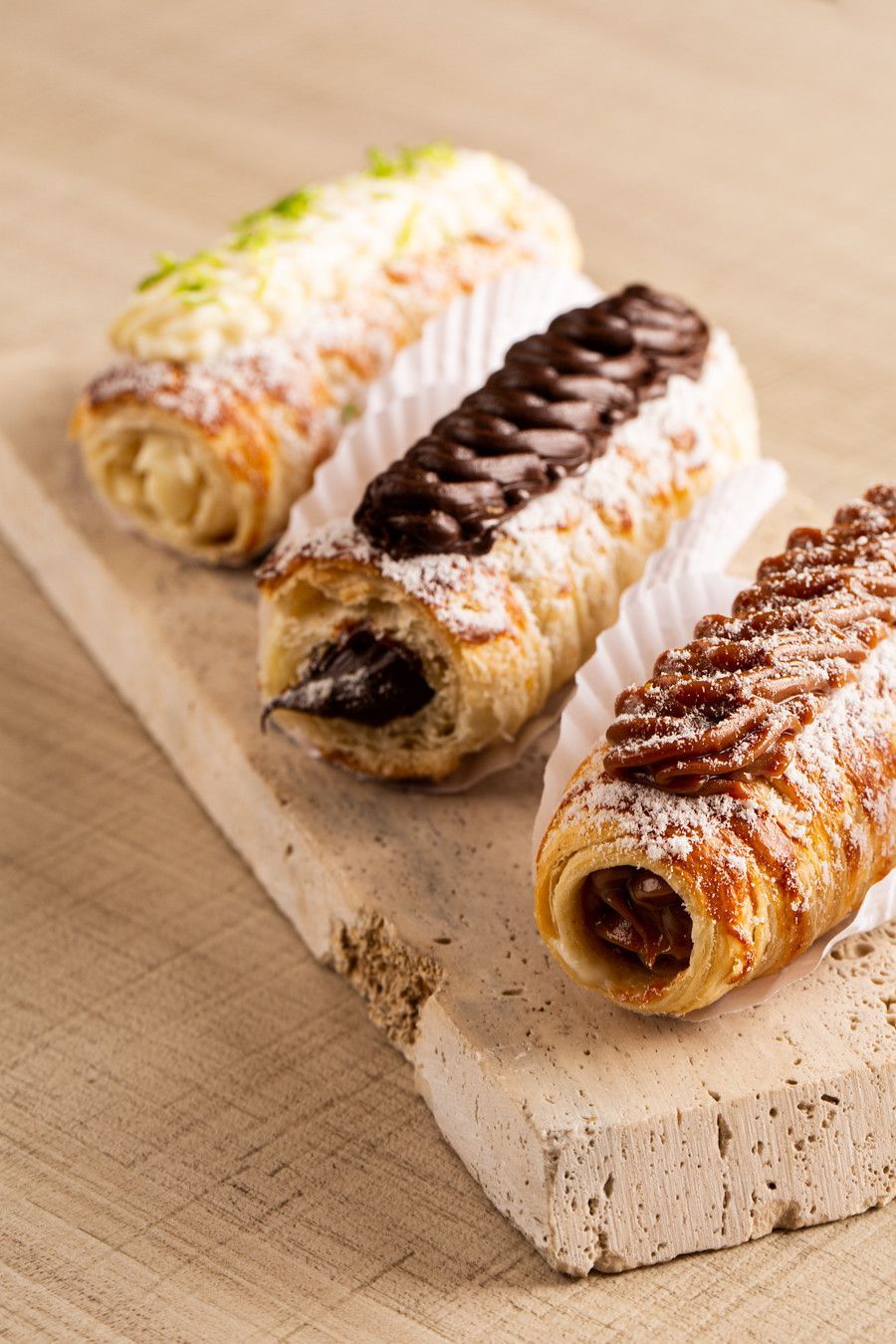 Três cannoli dispostos em uma tábua de madeira, cobertos com glacê branco, de chocolate e canela.