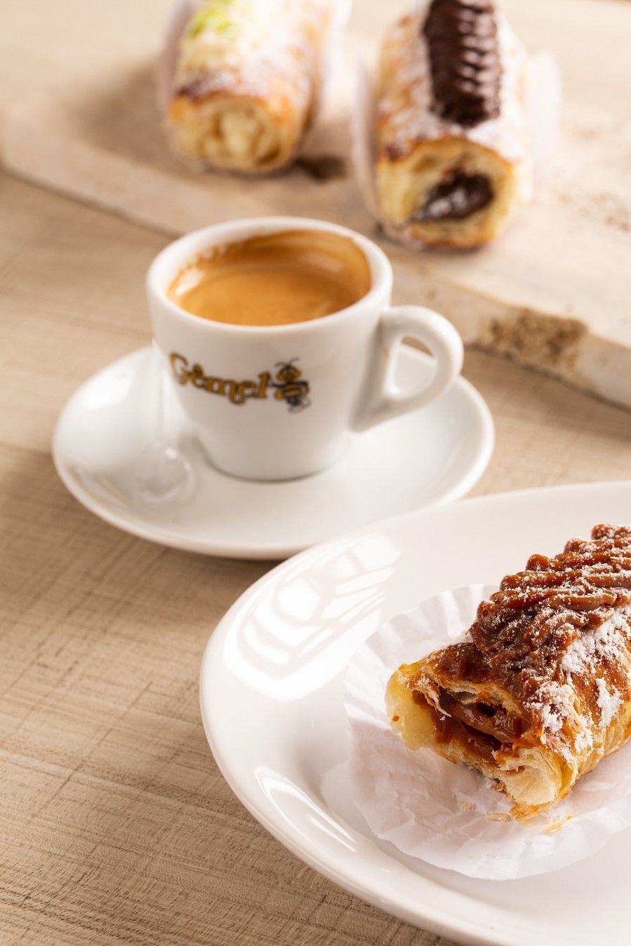 Xícara de café e doce em pratos brancos sobre uma mesa de madeira, com mais doces ao fundo.