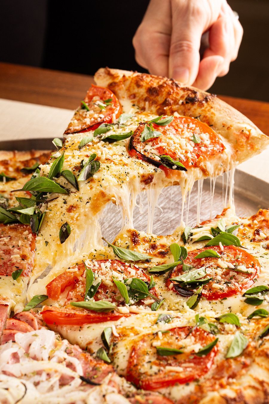 Mão agarrando uma fatia de pizza com tomate, manjericão e queijo derretido, estendendo-se da pizza.