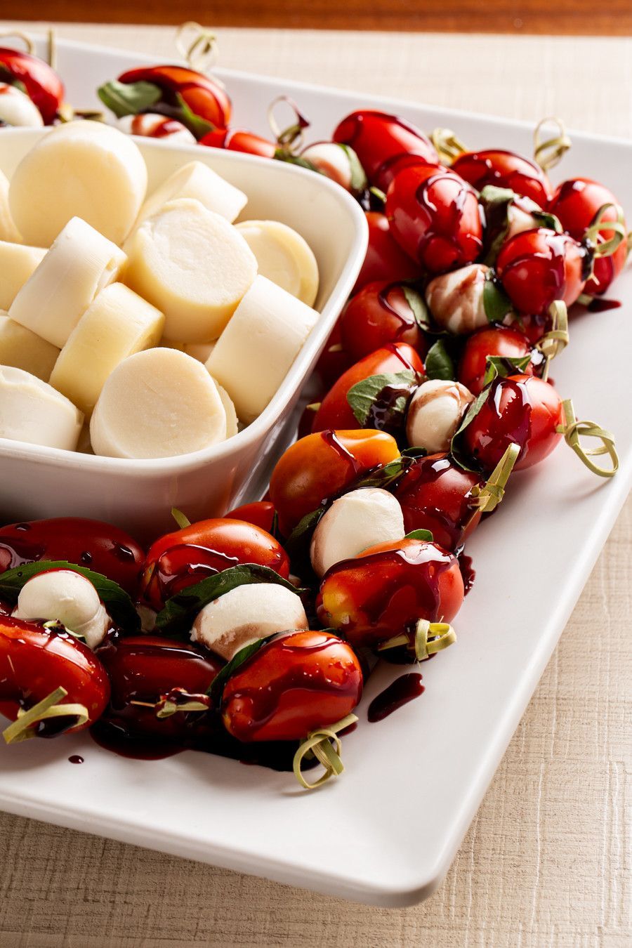 Espetos de tomates-cereja, bolinhas de mussarela e manjericão em um prato branco, acompanhados de uma tigela pequena com cubos de queijo.