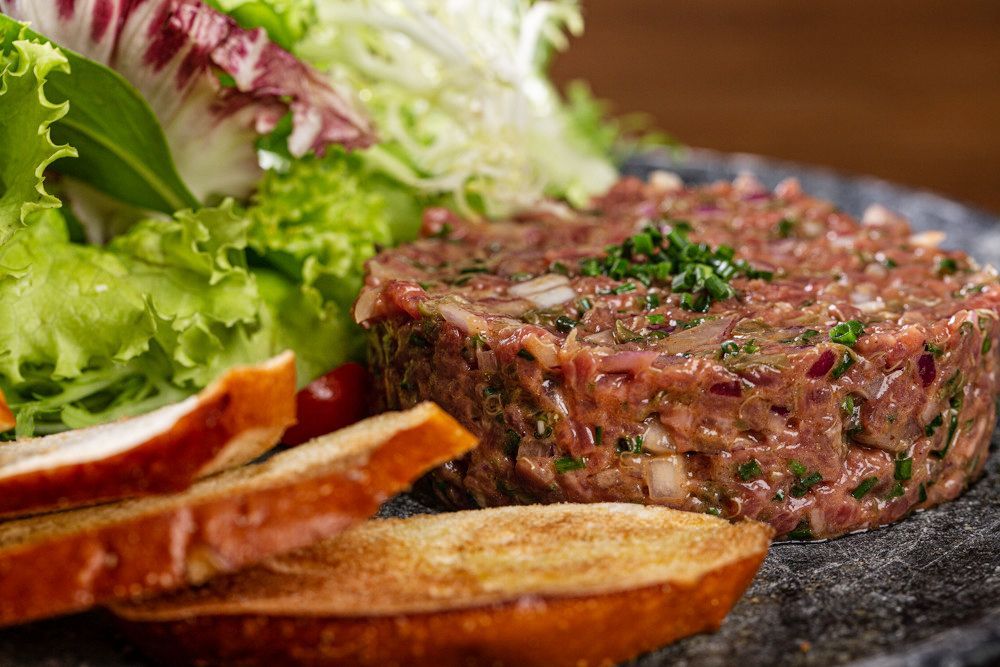 Steak tartare com pão torrado e salada verde em uma travessa de ardósia escura.