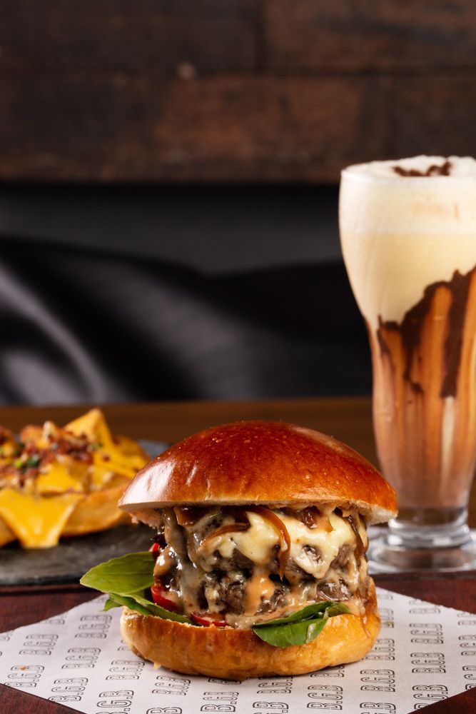 Cheeseburger com molho derretido ao lado de nachos e uma bebida alta e espumosa em uma mesa escura.
