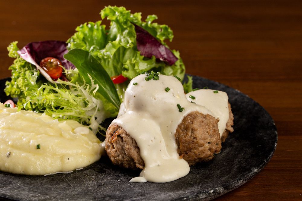 Almôndegas servidas em um prato escuro, cobertas com molho cremoso, purê de batatas e mix de folhas verdes.