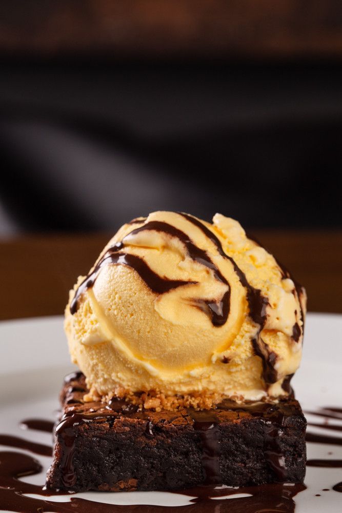Uma bola de sorvete de baunilha sobre um brownie, com calda de chocolate derretida, em um prato branco.