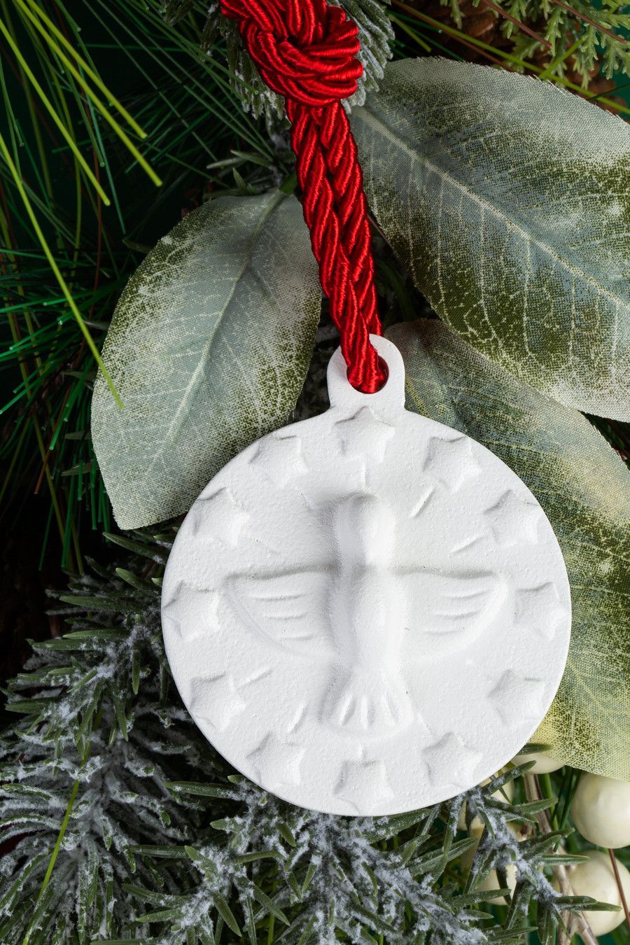 Ornamento medalhão redondo branco com figura em relevo, pendurado por um cordão vermelho sobre fundo verde.