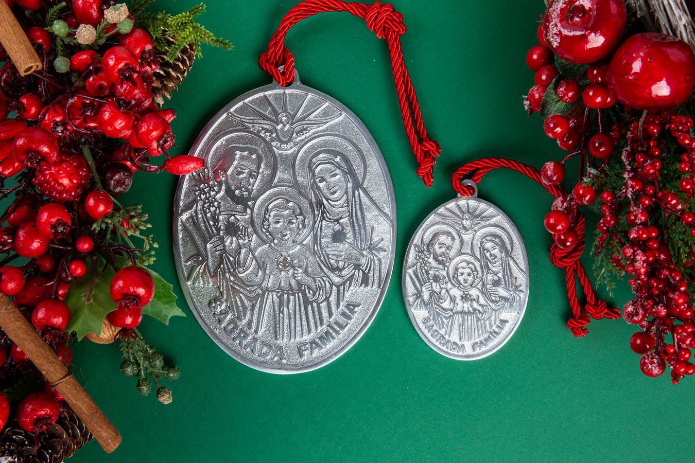 Dois medalhões religiosos de prata com cordões vermelhos sobre fundo verde, emoldurados por decorações natalinas vermelhas.