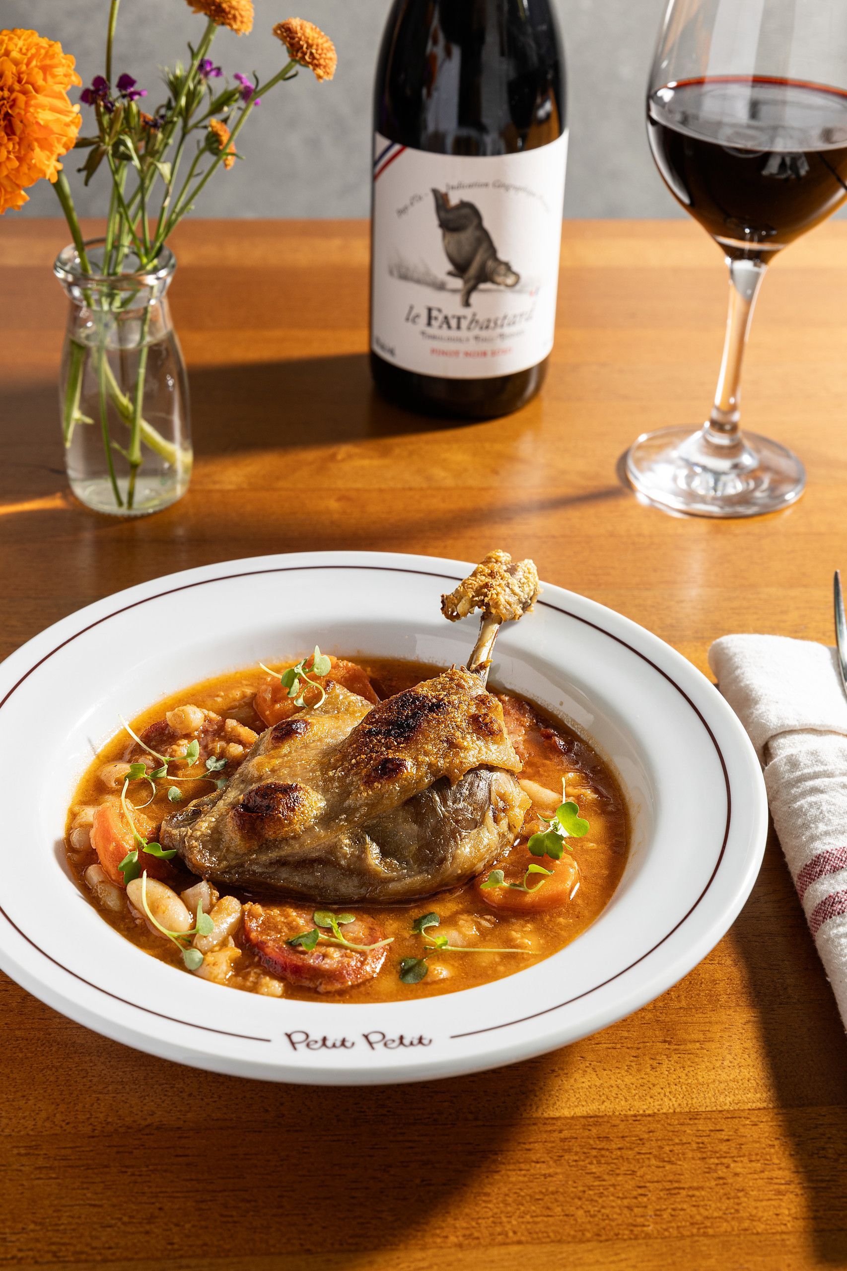 Peixe assado em uma tigela com caldo, flores, uma garrafa de vinho e um copo sobre uma mesa de madeira.