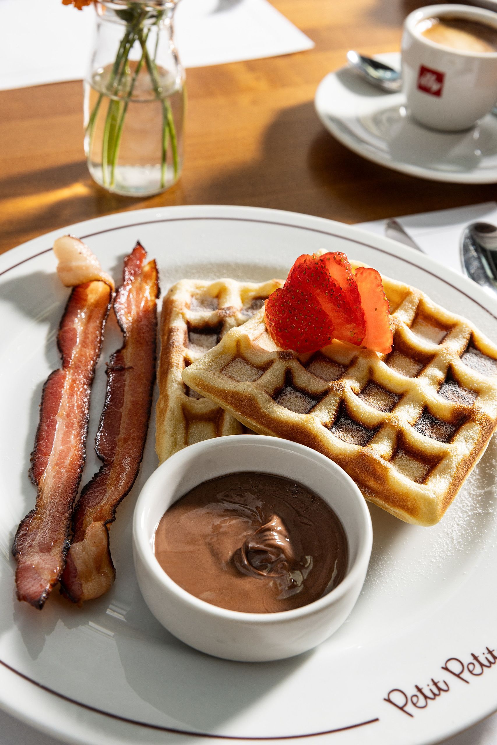 Waffle com morangos, bacon e calda de chocolate em um prato branco ao lado de uma xícara de café em uma mesa de cafeteria.