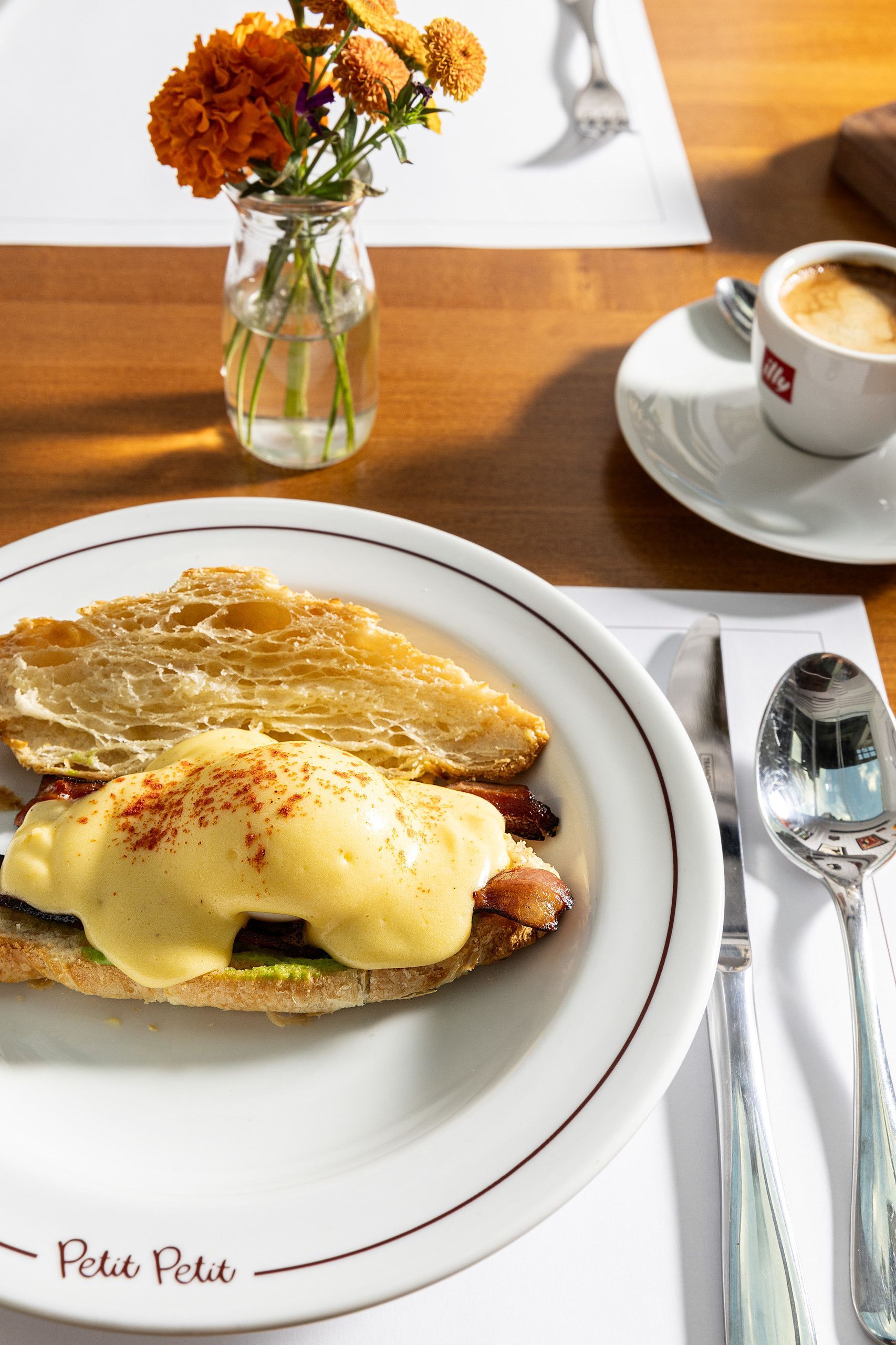 Ovos Benedict em um prato branco com café e flores sobre uma mesa de madeira.