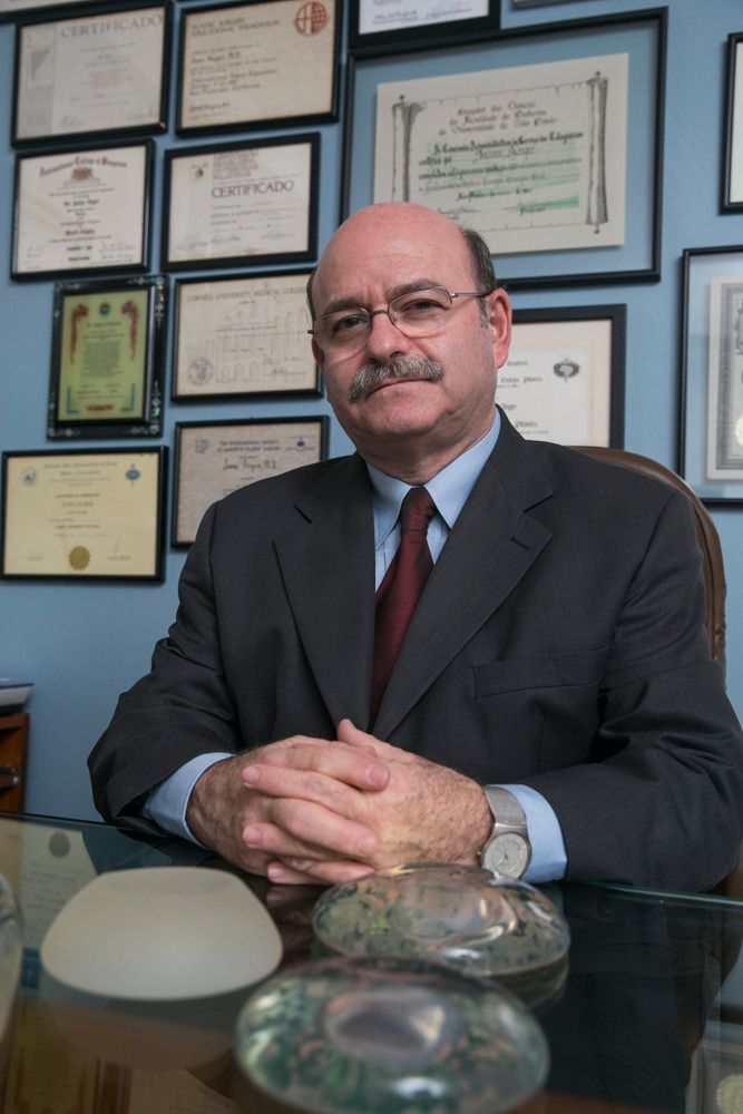 Homem de terno sentado em uma mesa com certificados emoldurados na parede atrás dele.