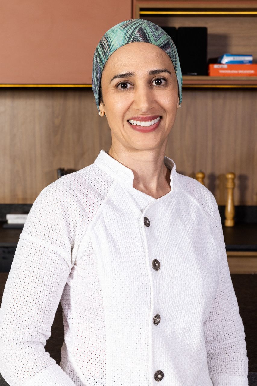 Pessoa sorridente vestindo blusa branca de botões e boné estampado, em pé em uma cozinha com prateleiras de madeira.