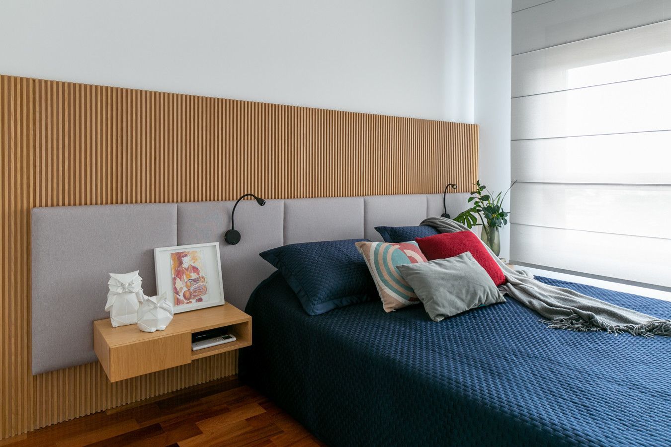 Quarto moderno com cama azul-escura, parede com ripas de madeira e mesa de cabeceira junto a uma janela bem iluminada.