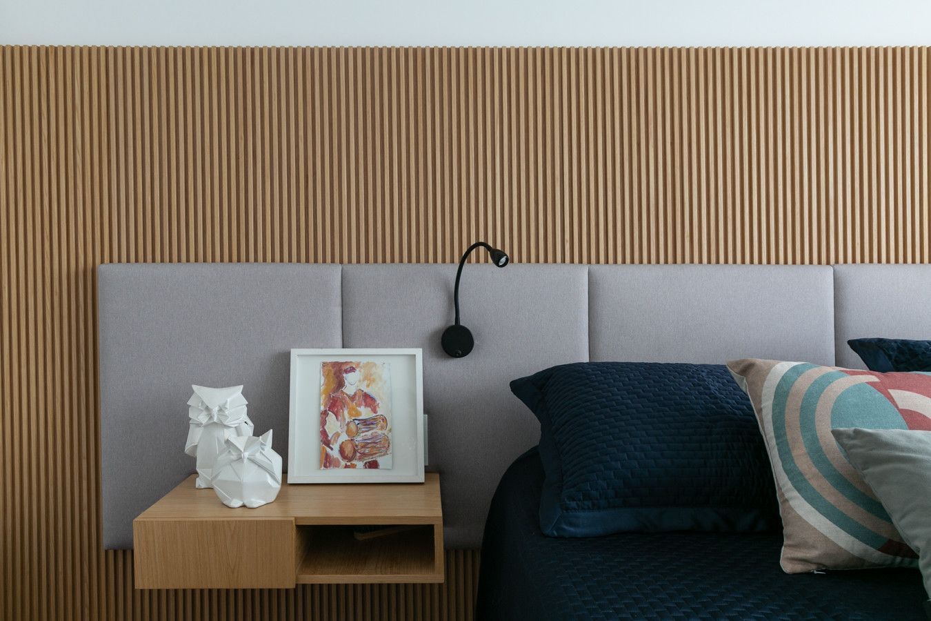 Quarto moderno com cabeceira cinza, mesa de cabeceira, abajur, quadro decorativo e roupa de cama azul-escura.