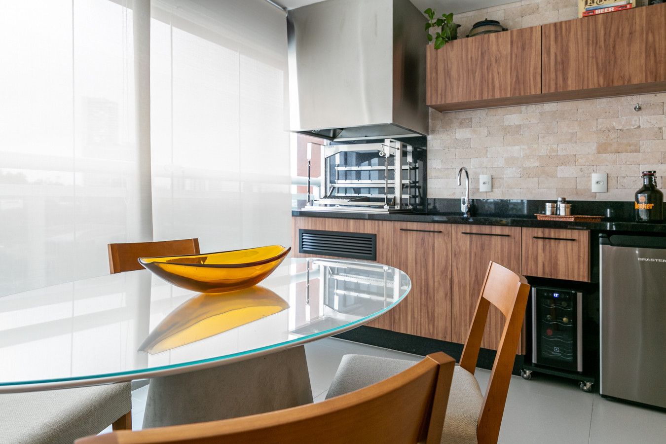 Cozinha moderna com mesa de jantar de vidro, cadeiras de madeira e uma fruteira junto a uma janela ensolarada.