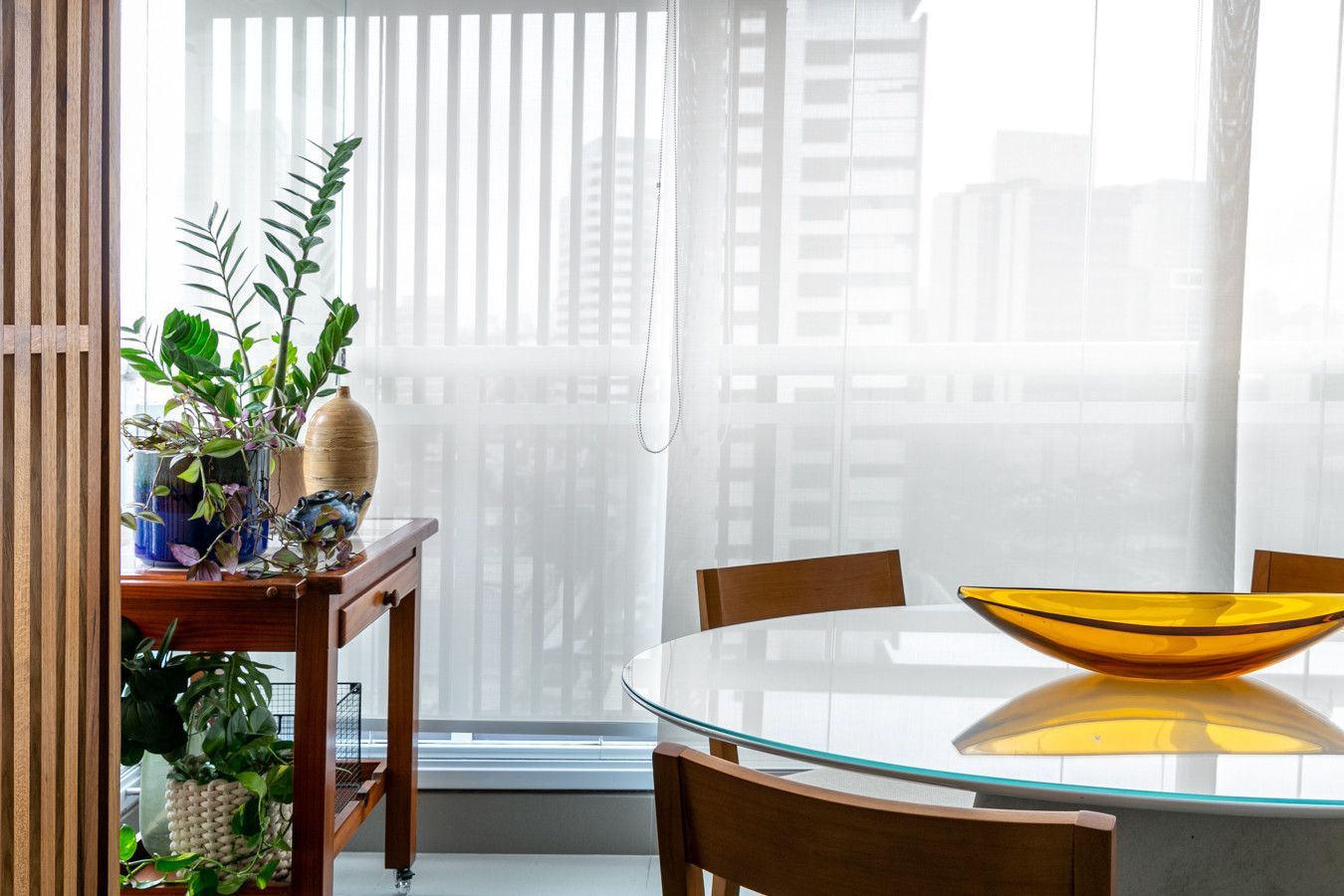 Sala de jantar iluminada com mesa de vidro, cadeiras de madeira, vasos de plantas e cortinas translúcidas junto à janela.