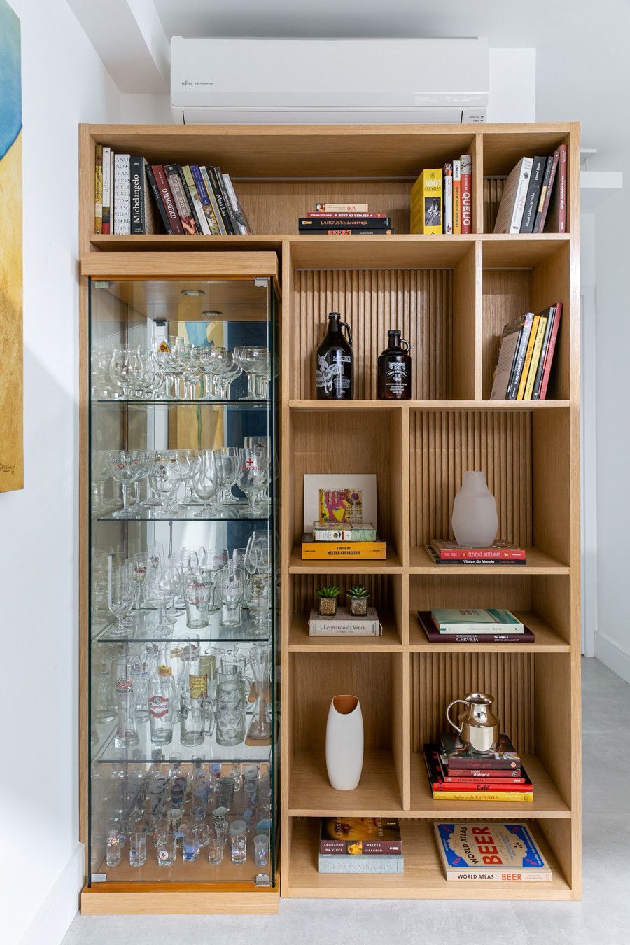 Vitrine de madeira com copos, livros e objetos decorativos em um ambiente bem iluminado.