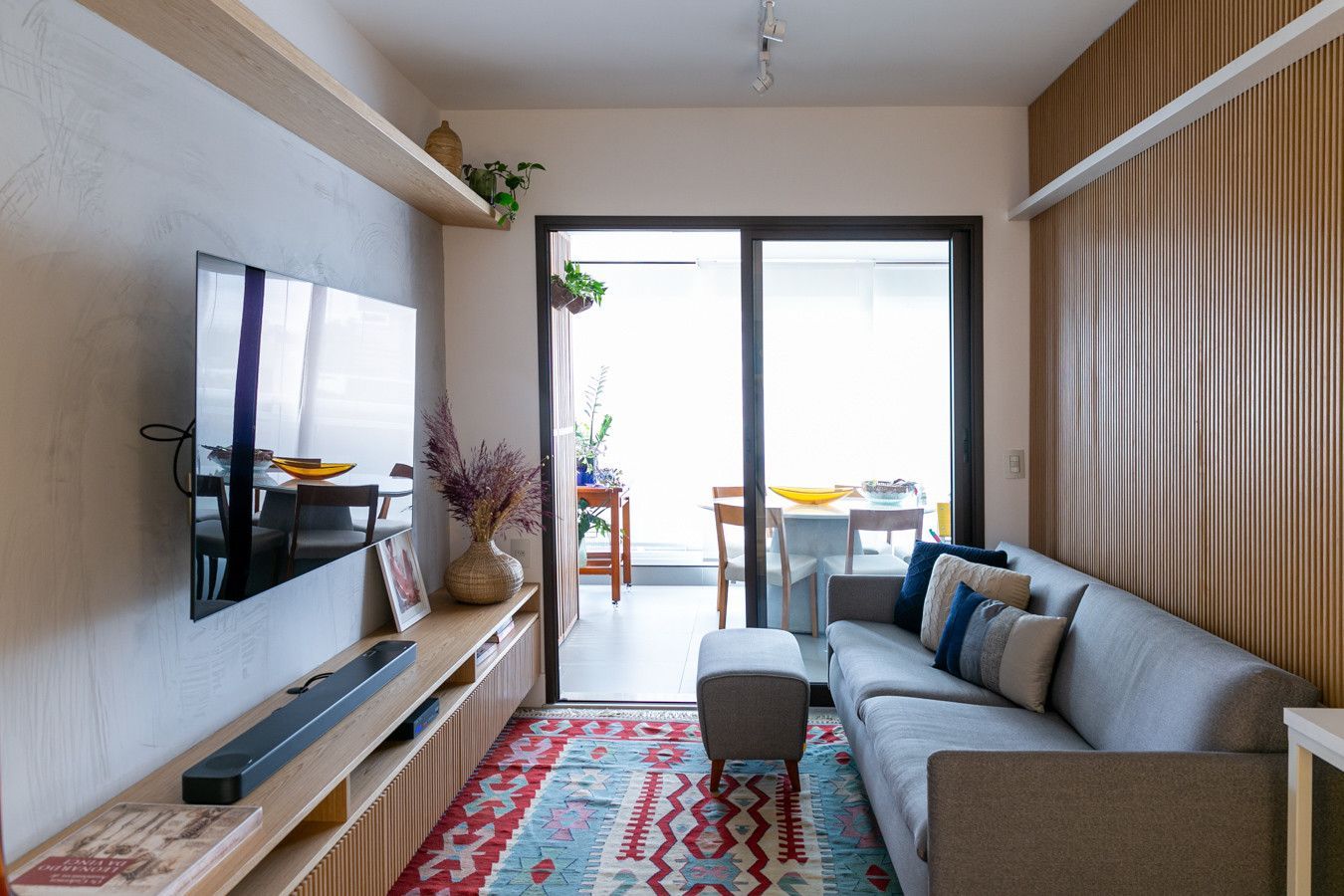 Sala de estar moderna com sofá cinza, TV de parede, tapete estampado e portas de sacada que permitem a entrada de luz natural.