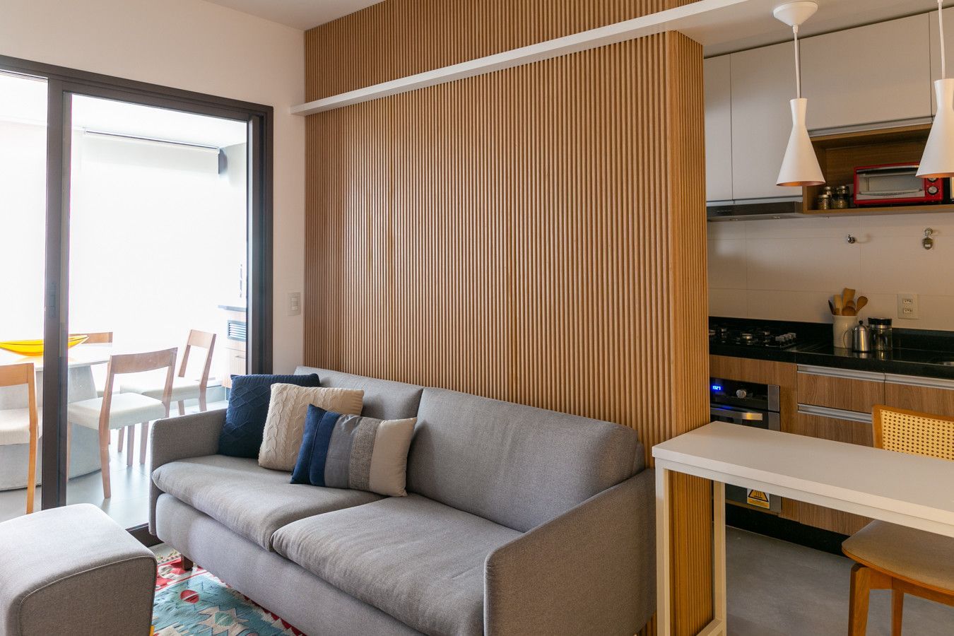 Sala de estar moderna com sofá cinza, cortinas listradas e cozinha integrada com janela para a varanda.