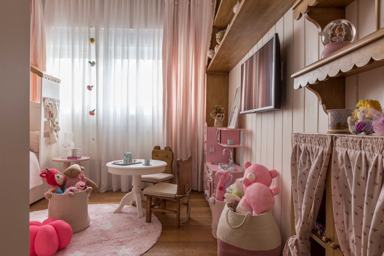 Sala de jogos infantil rosa com bichinhos de pelúcia, mesinha e uma janela iluminada com cortinas translúcidas.