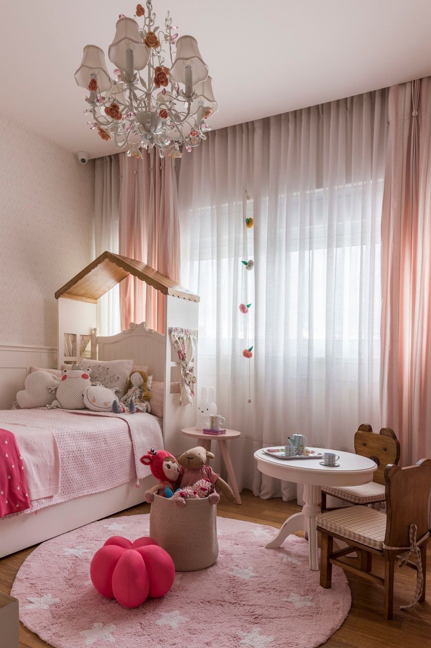 Quarto infantil rosa com cama de dossel, lustre e mesa de atividades junto a uma janela com cortina.