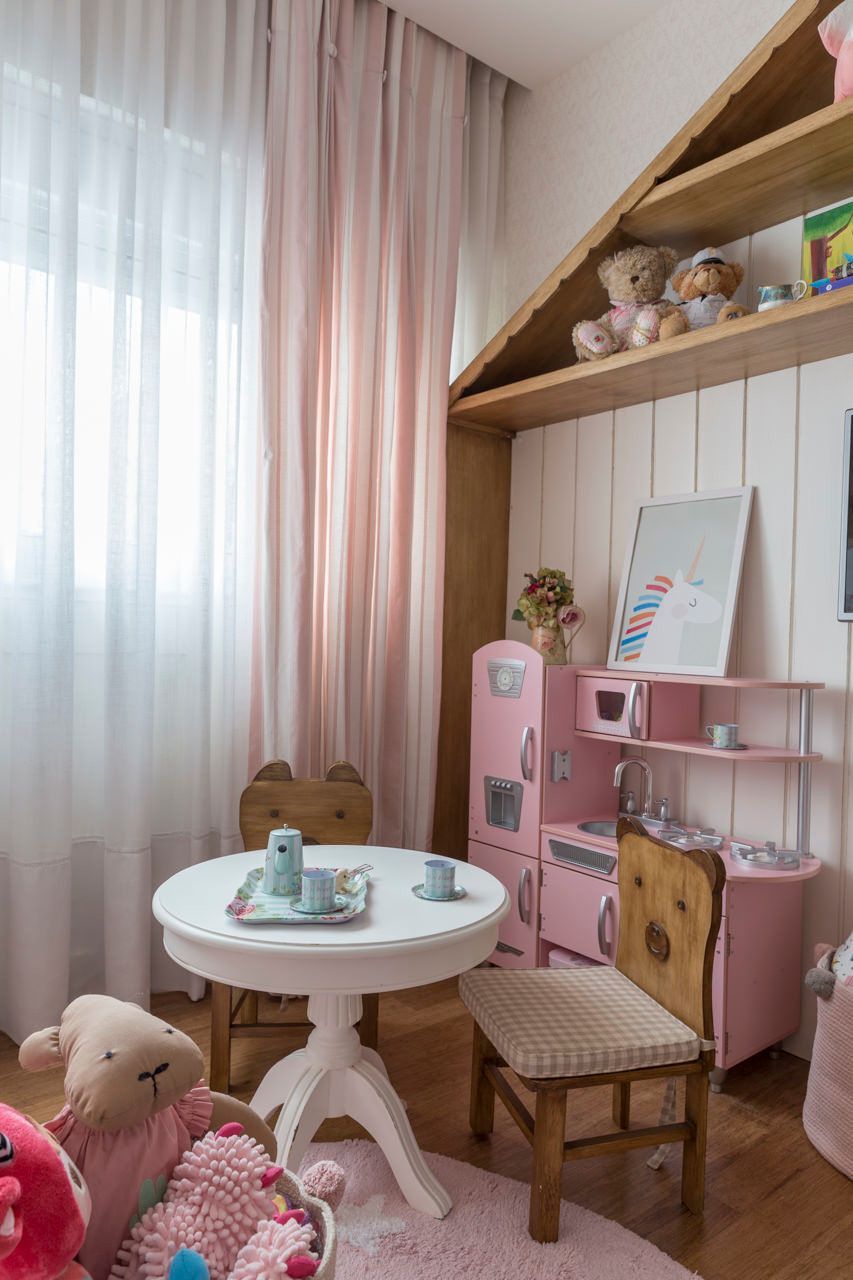 Quarto infantil rosa com uma mesa redonda, bichos de pelúcia e uma escrivaninha pequena perto da janela.