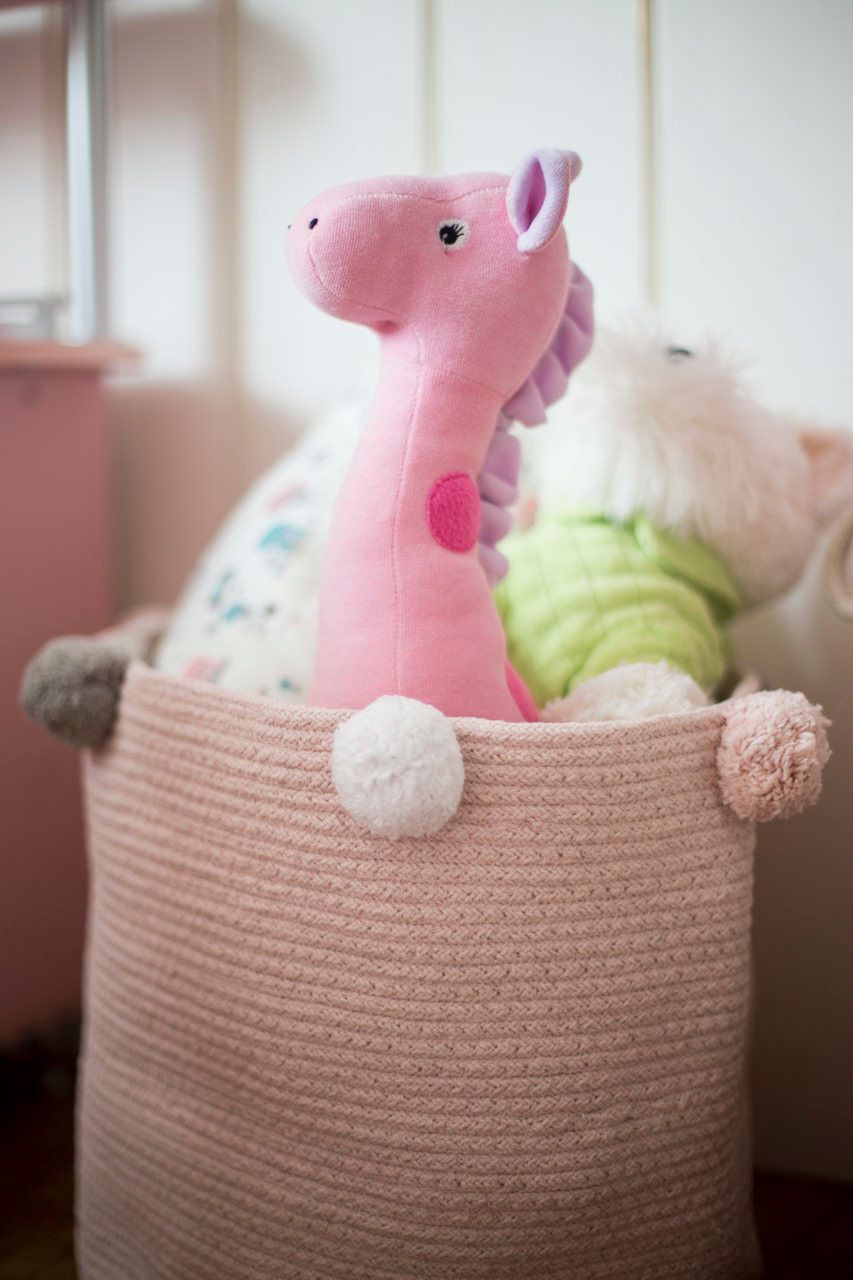 Dinossauro de pelúcia rosa em uma cesta de tricô com um brinquedo de pelúcia branco em um quarto de bebê com iluminação suave.