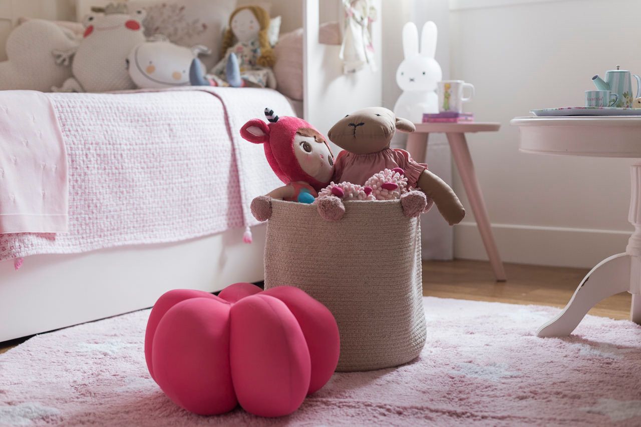 Quarto infantil com tapete rosa, cesto de brinquedos, bichinhos de pelúcia e um abajur branco em forma de coelho sobre um banquinho.