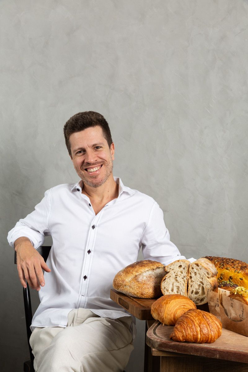 Homem sentado ao lado de uma mesa com diversos tipos de pão, sorrindo, vestindo camisa branca contra um fundo cinza.