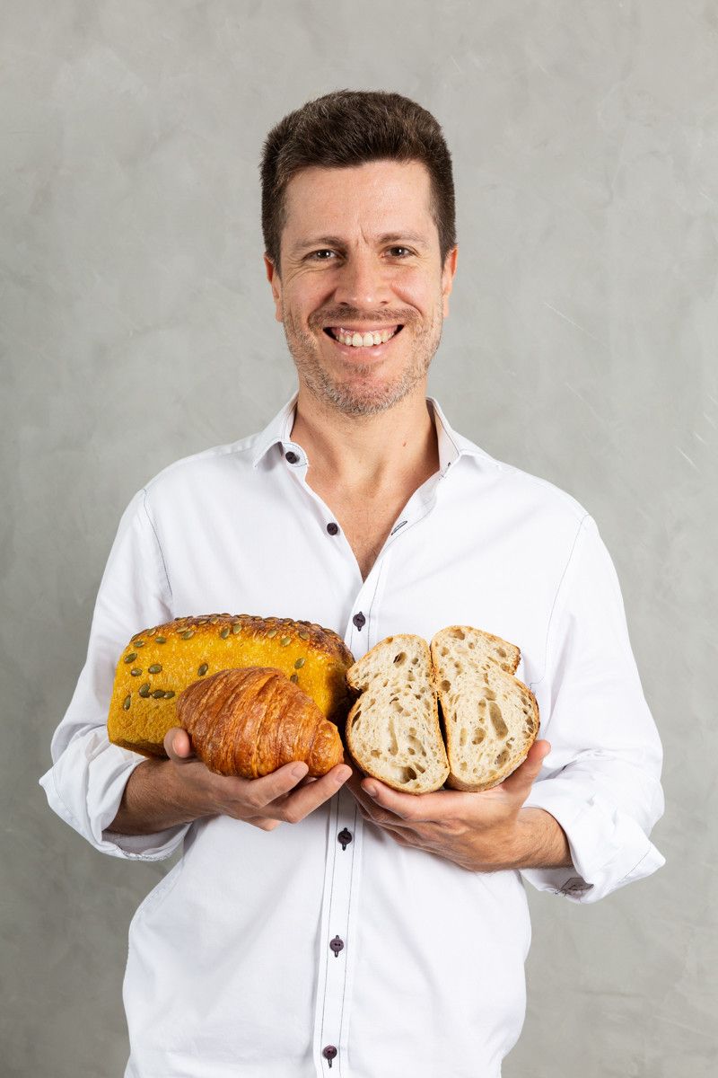 Homem segurando dois pães, sorrindo contra um fundo cinza.