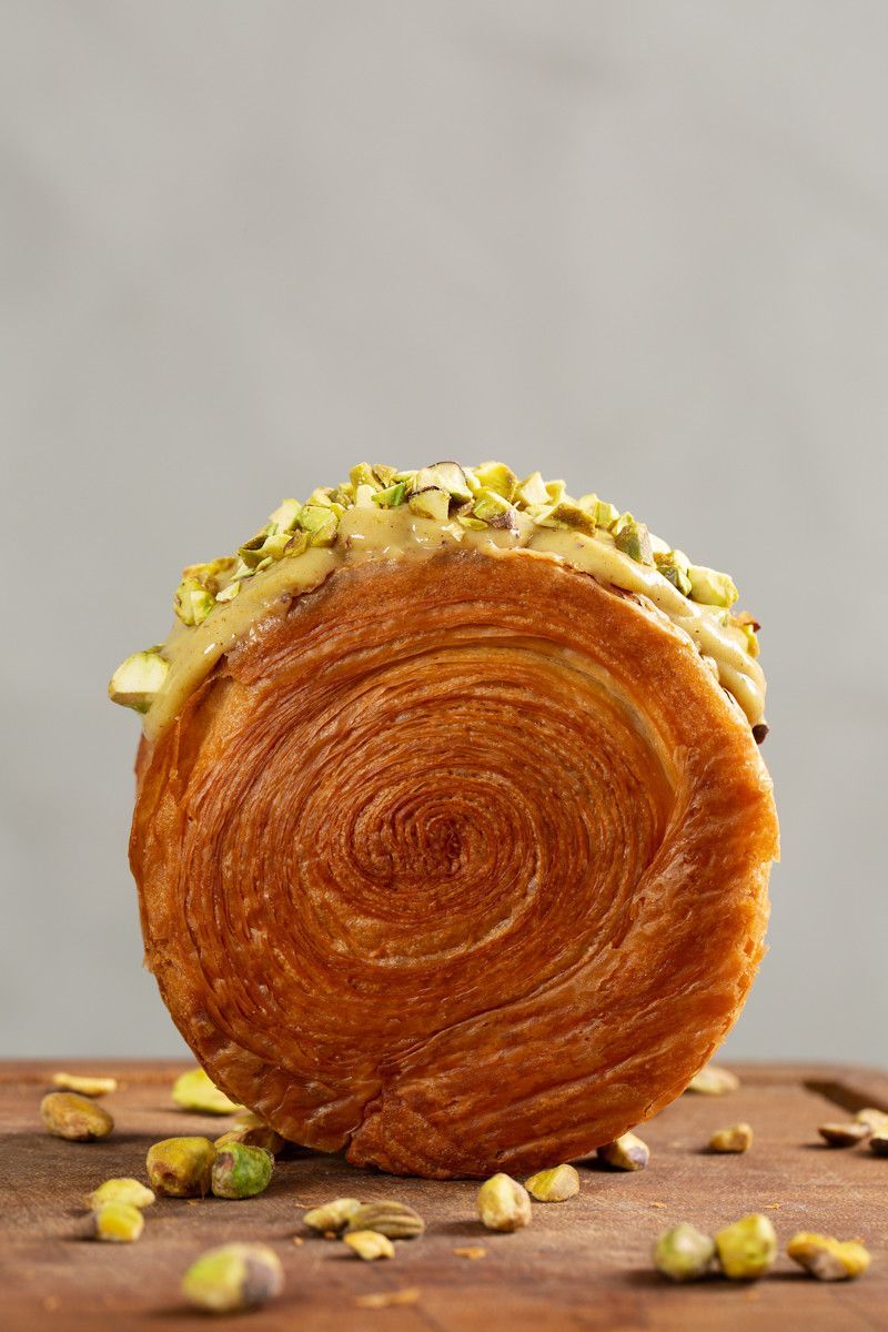 Pastel redondo coberto com pistache, disposto sobre uma superfície de madeira com nozes espalhadas, em um fundo cinza.
