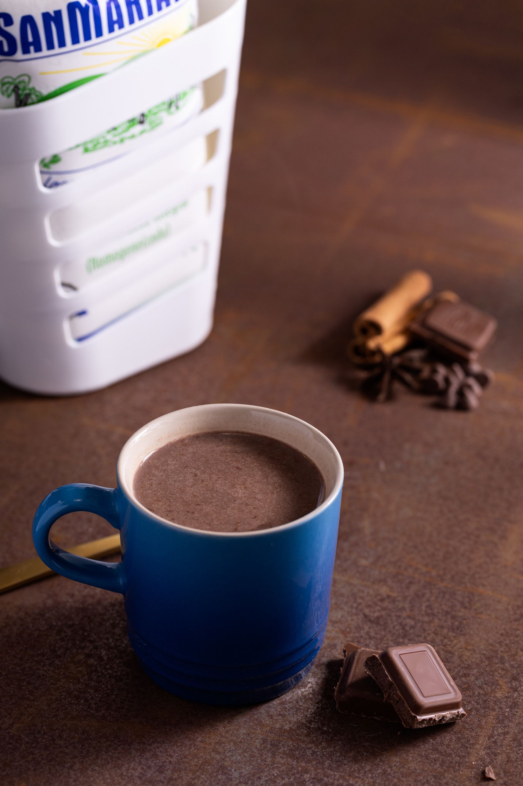Caneca azul de chocolate quente com pedaços de chocolate ao lado de um recipiente branco sobre uma mesa marrom.