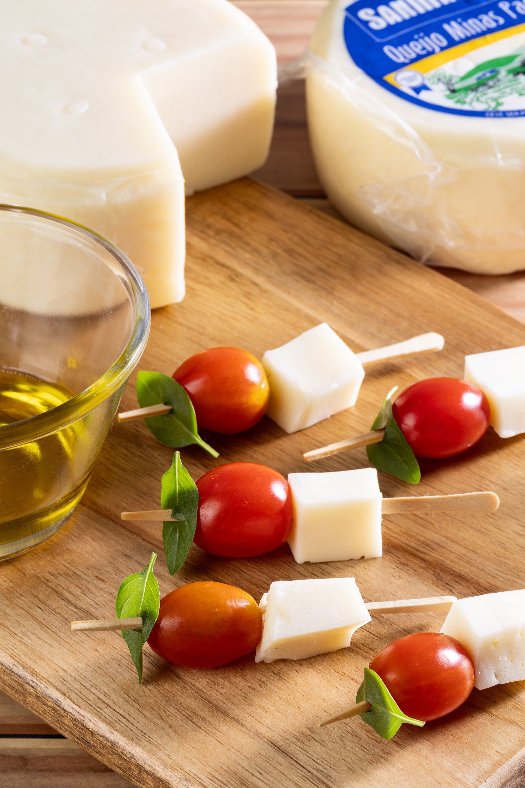 Espetinhos de tomate-cereja, cubos de mussarela e manjericão ao lado de azeite e queijo em uma tábua de madeira.
