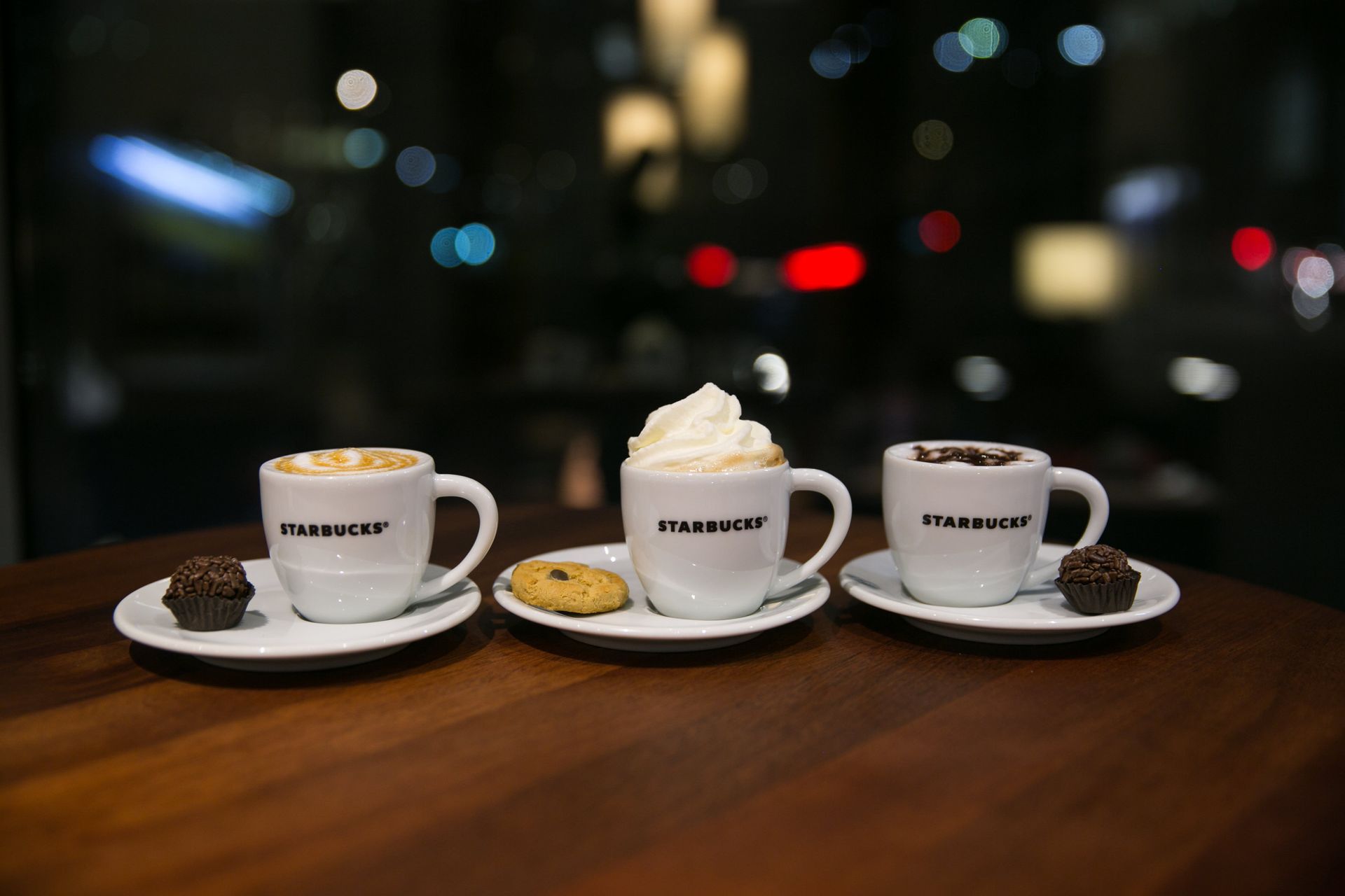 Três xícaras de café e sobremesas em pratos sobre uma mesa de madeira em um café à noite.
