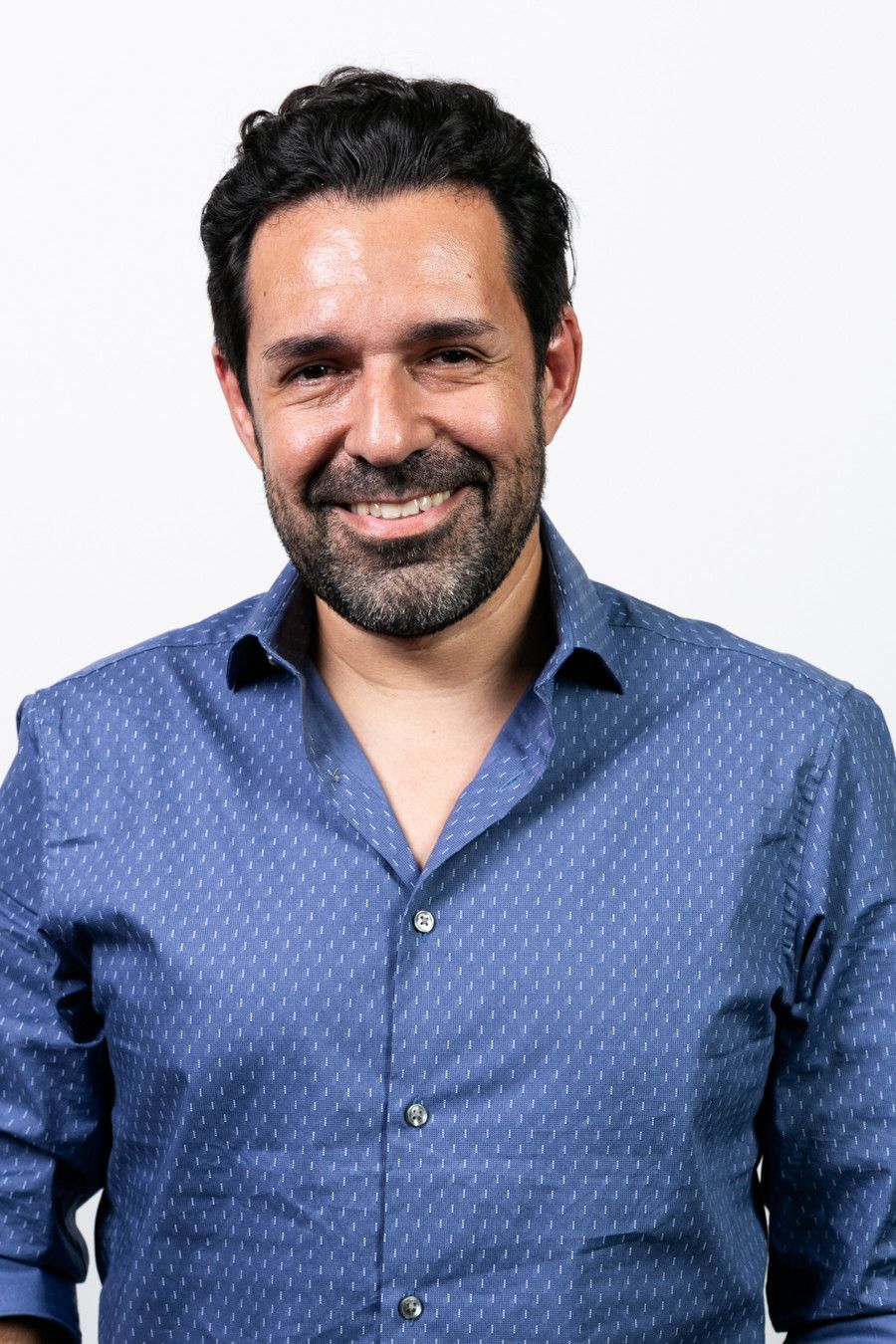 Homem sorridente com camisa azul estampada em pé contra um fundo branco.