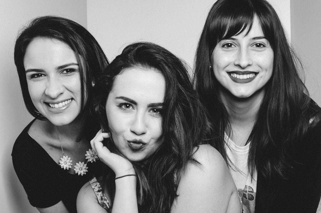 Três mulheres posando bem próximas uma da outra, sorrindo para a câmera em um retrato em preto e branco.