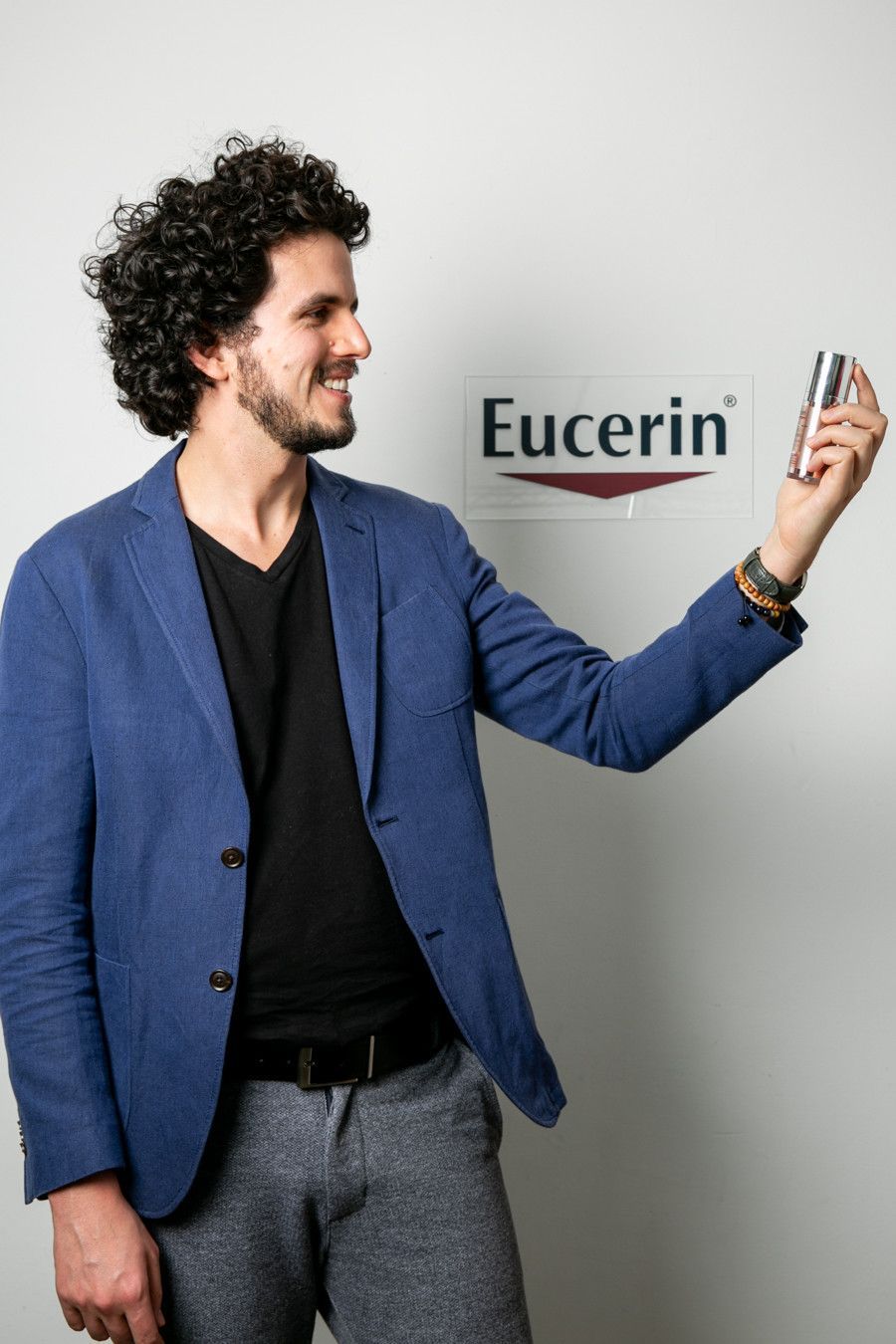 Homem de blazer azul segurando um produto Eucerin e sorrindo contra um fundo cinza.